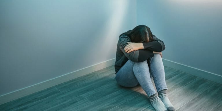 woman feeling depressed sitting in corner of empty room with arms crossed and head down on knees