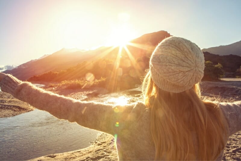 woman wearing gray knit hat looking out toward bright sun with river and mountains
