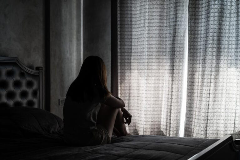 Sad woman sitting on a bed in a dimly lit room, reflecting emotional distress or depression