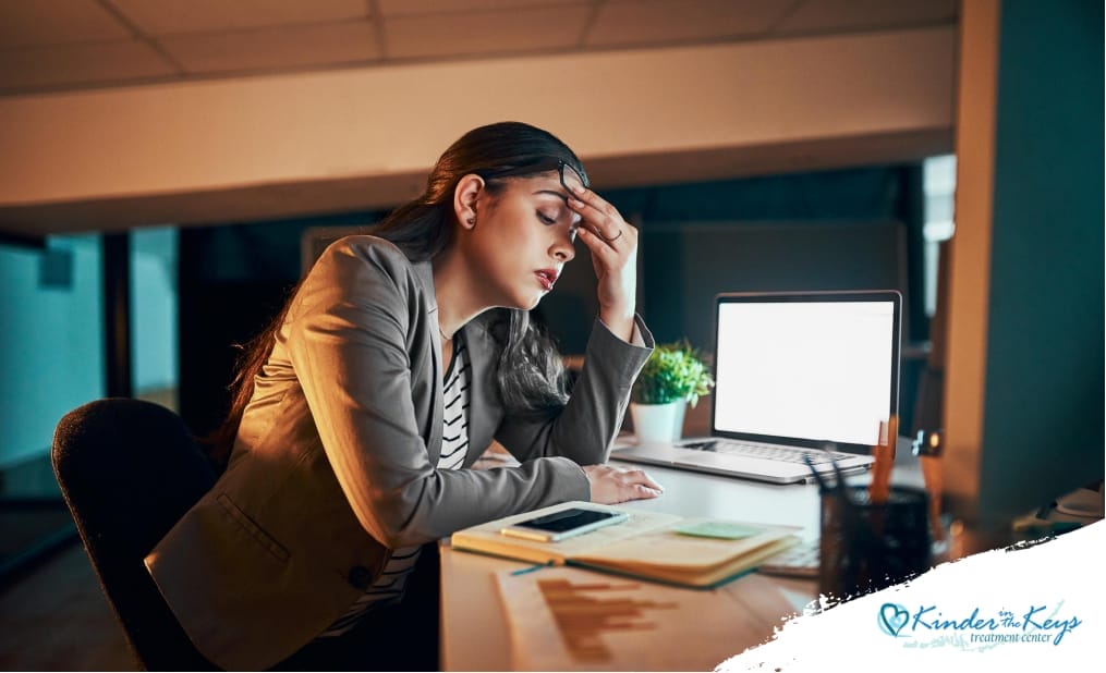 Close-up of a woman sitting at her desk, visibly stressed, with her hand on her forehead, surrounded by papers and a laptop, indicating pressure and the possibility of a mental breakdown.