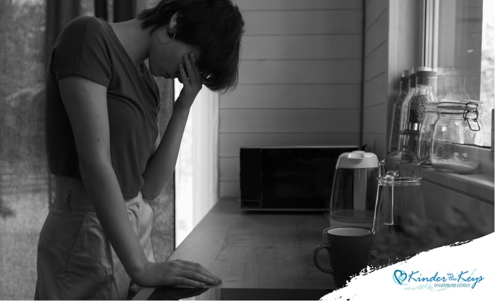 A woman standing at the kitchen sink with her head bowed, looking overwhelmed and distressed, showing signs of a nervous breakdown, surrounded by dishes and a cluttered countertop.