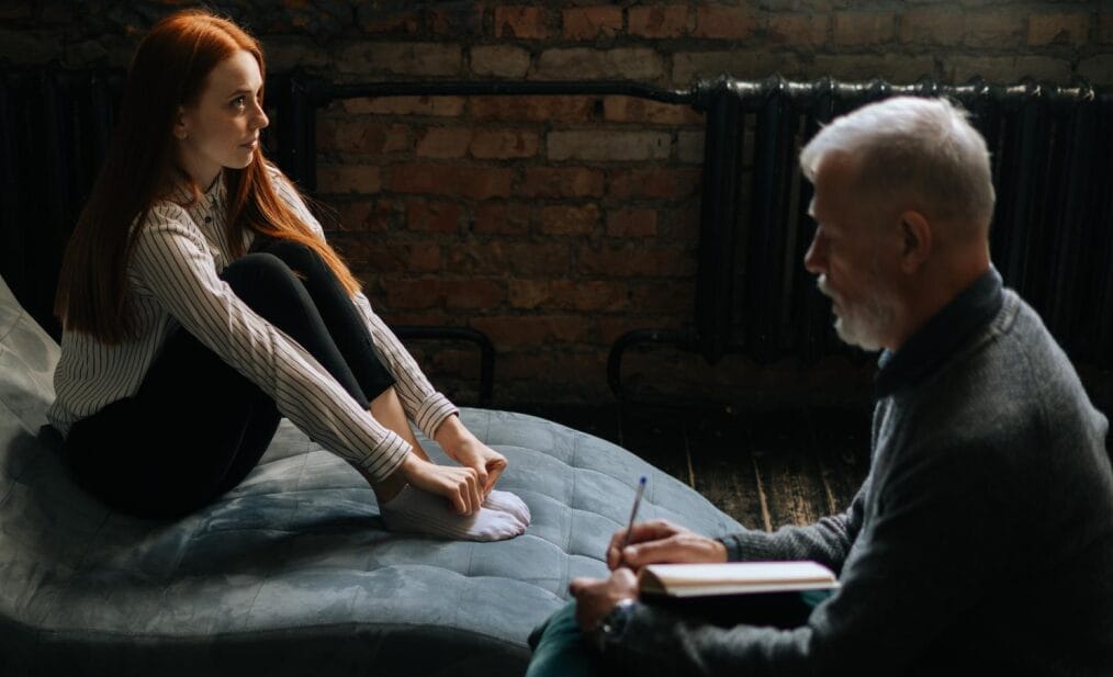 Professional gray-haired man psychologist consulting patient during psychological consultation in cozy dark office room. Bearded mature psychotherapist taking notes and giving advices to patient.